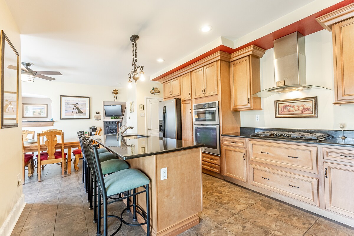 A spacious gourmet kitchen is featured with wooden cabinetry and modern appliances. A large island with bar seating offers a gathering space. An adjacent dining area is visible, showcasing a wooden dining table and chairs, enhancing the home’s functionality.