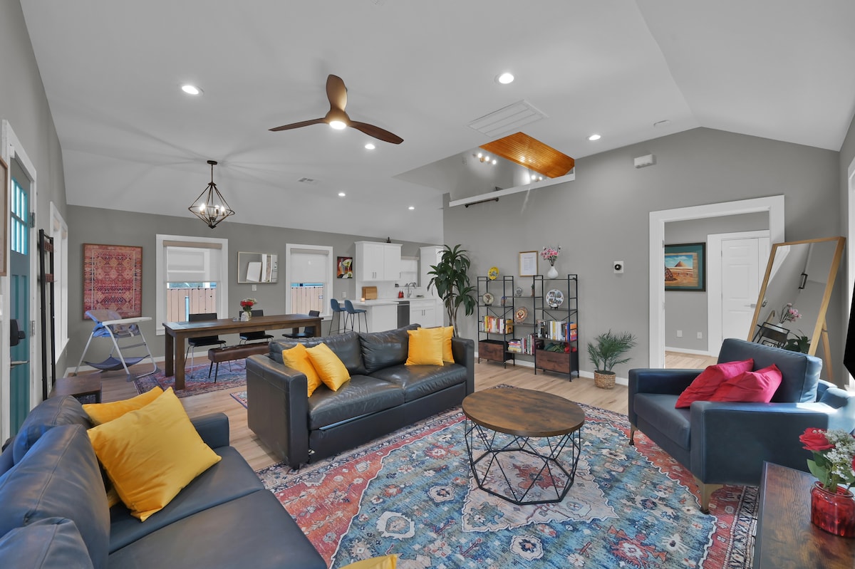The living room features two dark sofas adorned with yellow pillows, complemented by a round wooden coffee table. A colorful area rug adds warmth to the space. Large windows allow natural light to illuminate the room, which includes a ceiling fan and modern lighting fixtures.