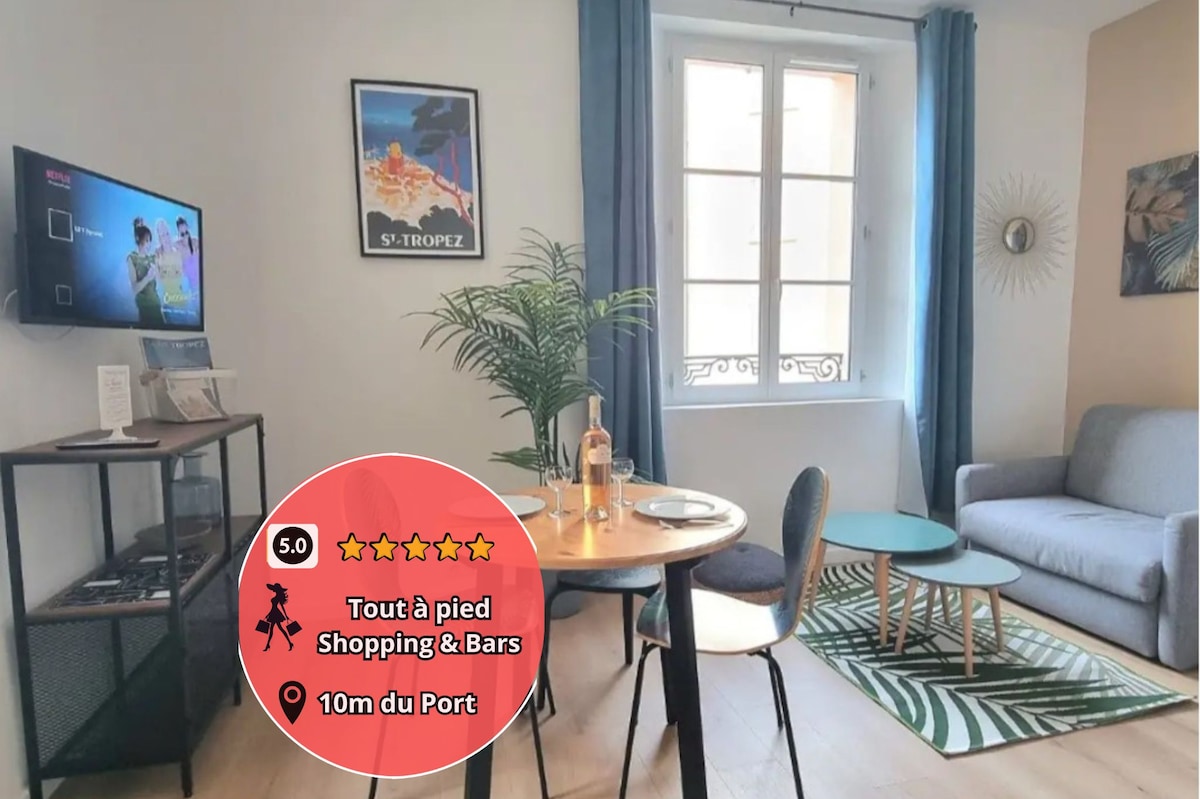 The cozy living area features a gray sofa and a round dining table with two chairs. A flat-screen TV is mounted on the wall. Natural light streams through the window, brightening the space adorned with a decorative plant.