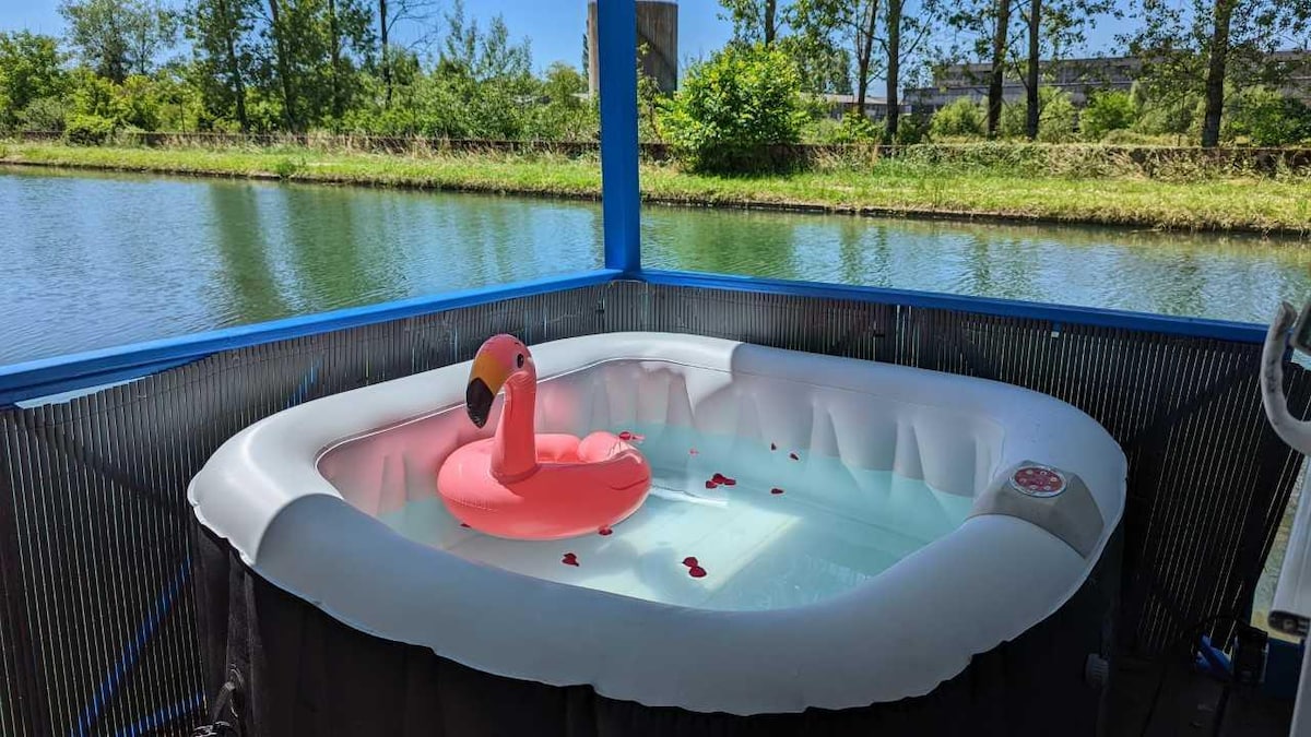 A private terrace features a jacuzzi filled with water, accompanied by a floating flamingo toy. The surrounding area is marked by lush greenery and a tranquil water view, creating a serene atmosphere for relaxation.