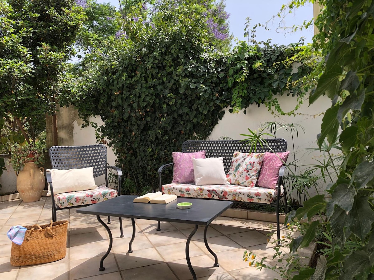 A serene outdoor space is surrounded by lush greenery. Two wrought iron benches with floral cushions face a small black table. A textured brown basket rests nearby, contributing to the tranquil atmosphere.