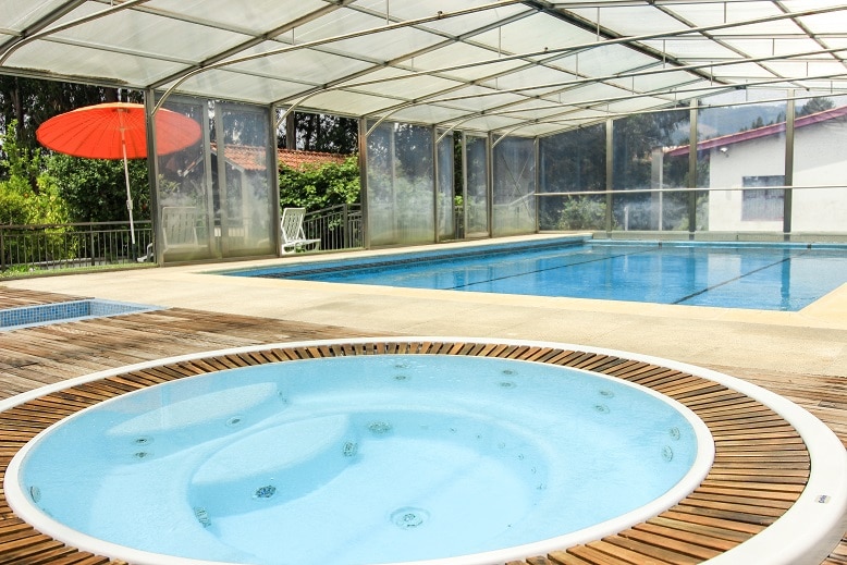A covered outdoor pool area is equipped with a spacious swimming pool and a separate jacuzzi. Wooden decking surrounds the jacuzzi, creating a relaxing space. Lush greenery is visible in the background, with a large umbrella providing shade in the area.