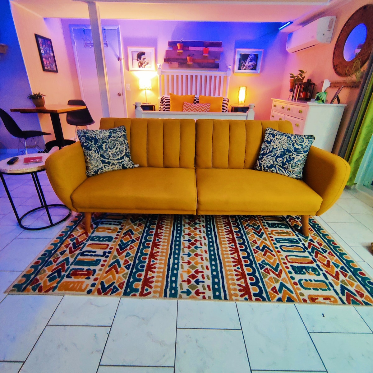 A modern living space is highlighted by a vibrant yellow sofa adorned with cushions, situated on a decorative area rug. Soft lighting enhances the inviting atmosphere, while a small table and two chairs, along with accent pieces on the walls, complete the cozy setup.