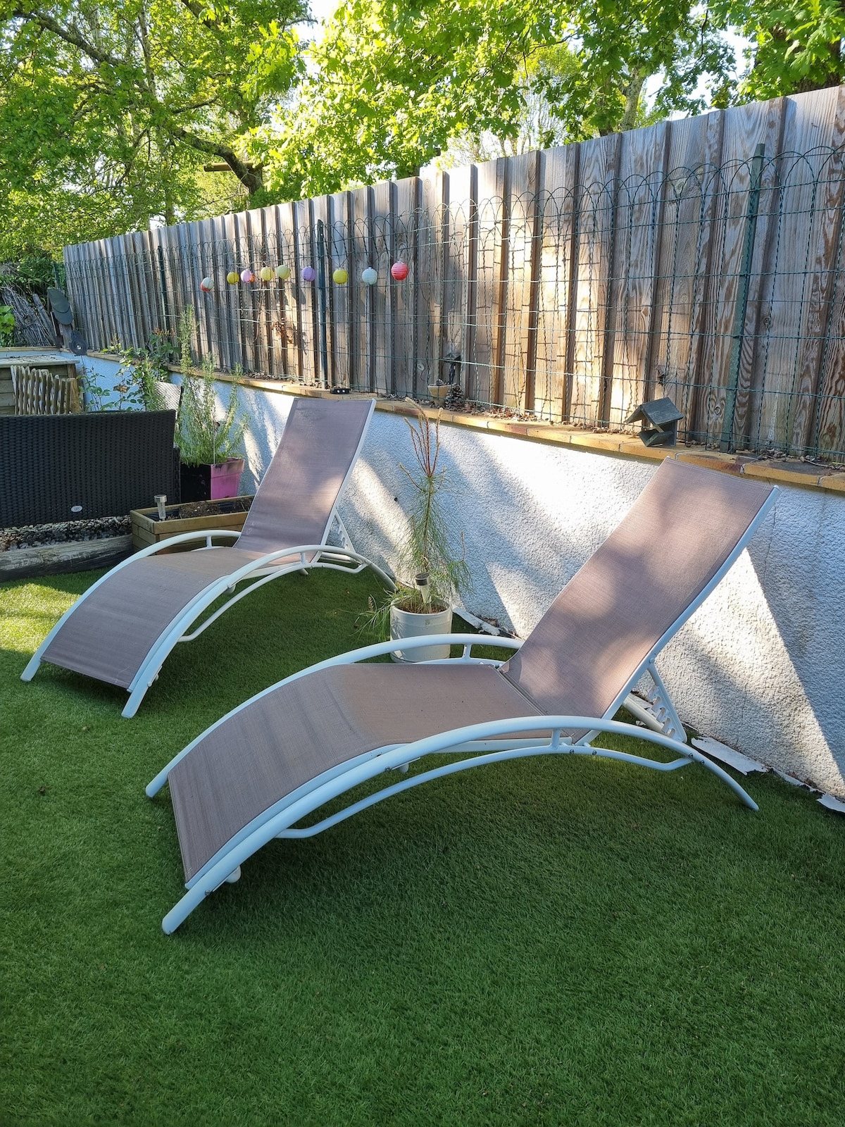 Two lounge chairs are positioned on a well-maintained grass surface, inviting relaxation outdoors. A wooden fence is visible in the background, adorned with decorative lighting and potted plants, enhancing the outdoor ambiance.