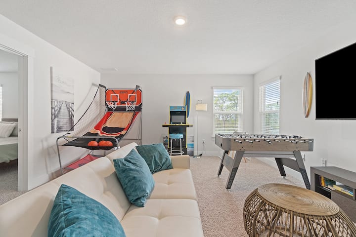 Game room featuring an indoor basketball arcade, retro-style video game machine, foosball table, and a cozy seating area with a white sofa and teal cushions. Decor includes a surfboard, wall-mounted TV, and natural light from multiple windows.