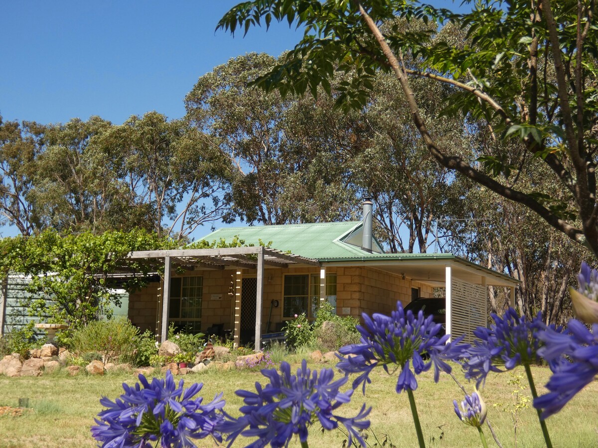 A charming cottage is nestled amidst native trees and vibrant flowers. The structure features a green roof and a welcoming porch, surrounded by natural greenery, creating a serene rural setting. The landscape includes scattered rocks and colorful plants, enhancing the outdoor experience.