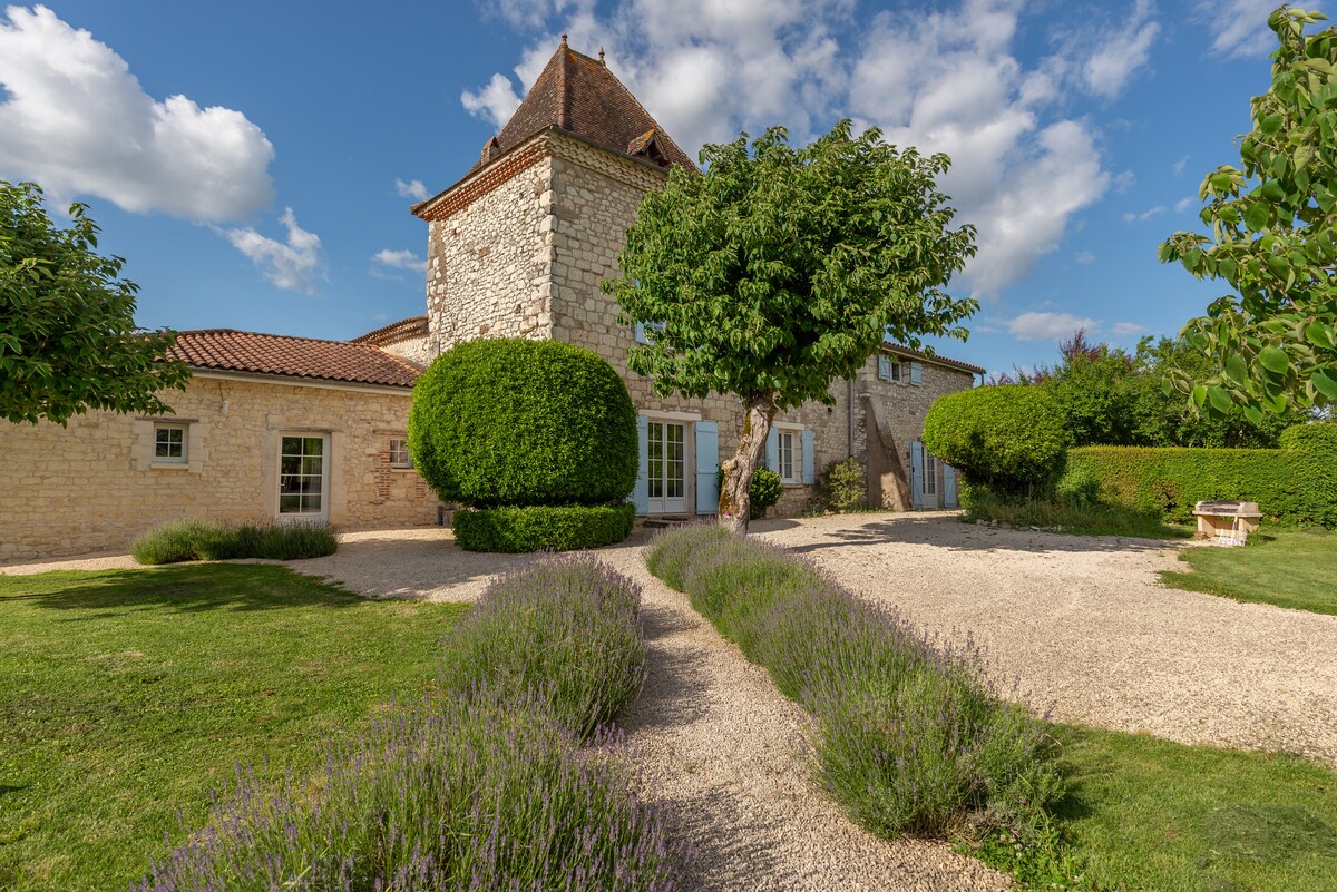 A charming stone house is surrounded by well-maintained gardens. Lavender plants line a gravel pathway leading to the entrance, bordered by lush green shrubs and trees. The sky above is dotted with fluffy clouds, enhancing the serene outdoor setting.