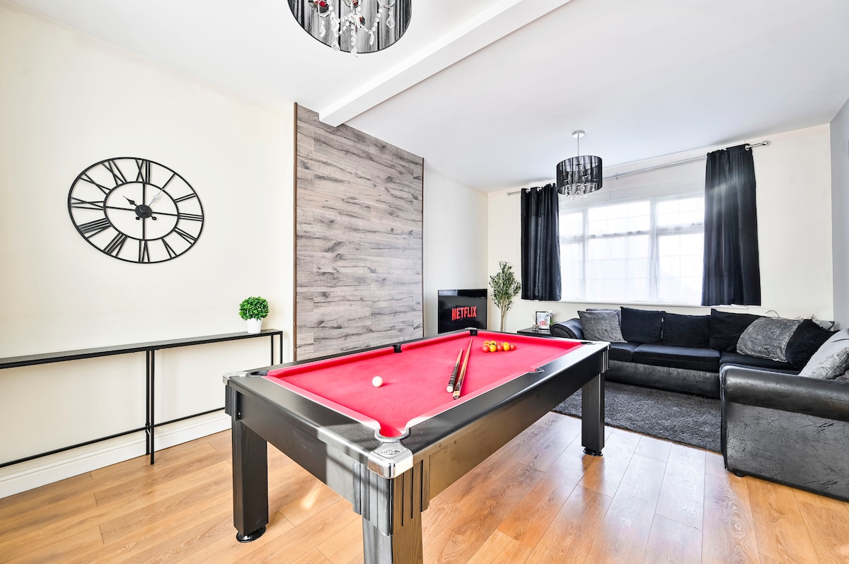 The living area features a red pool table at the forefront, surrounded by a stylish corner sofa and a television displaying streaming options. A large wall clock and modern light fixtures complement the warm wood flooring, creating a relaxed space for entertainment.
