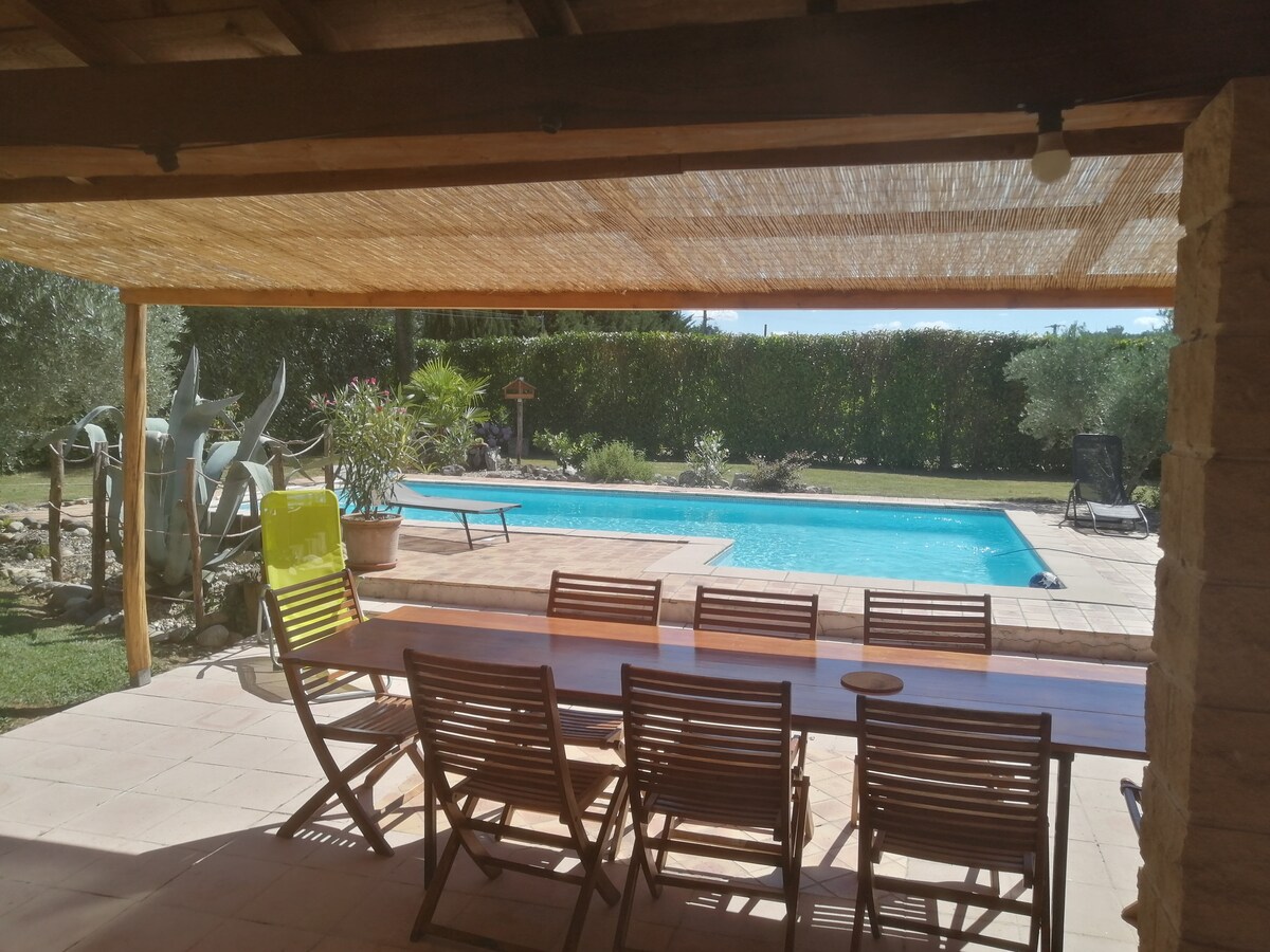 An outdoor terrace is presented, featuring a long wooden dining table surrounded by several folding chairs. Beyond, a swimming pool glistens under the sun, framed by greenery and a sunshade overhead. The tranquil setting is enhanced by potted plants and sculptures in the background.