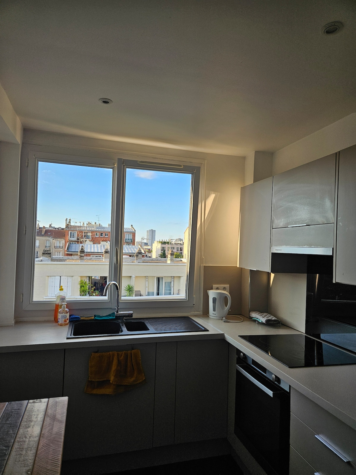 A bright kitchen features modern appliances and a large window overlooking the rooftops of the neighborhood. Natural light fills the space, highlighting the sleek cabinetry and the kitchen sink positioned beneath the window. A wooden table adds warmth to the modern design.
