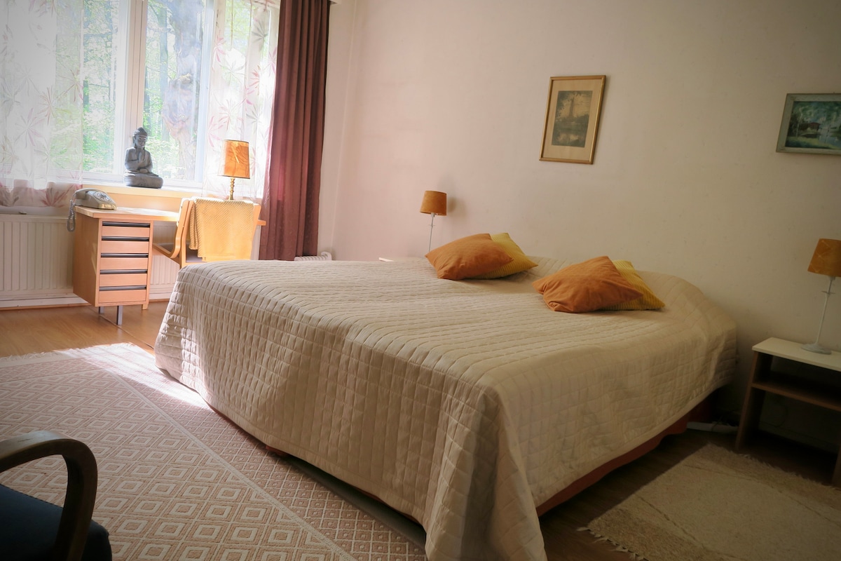 A spacious bedroom features a large bed covered with a light quilt. Two decorative cushions are placed on the bed. Natural light filters through sheer curtains, illuminating the warm-toned decor. A small desk and lamp are situated near the window, enhancing the tranquil ambiance.