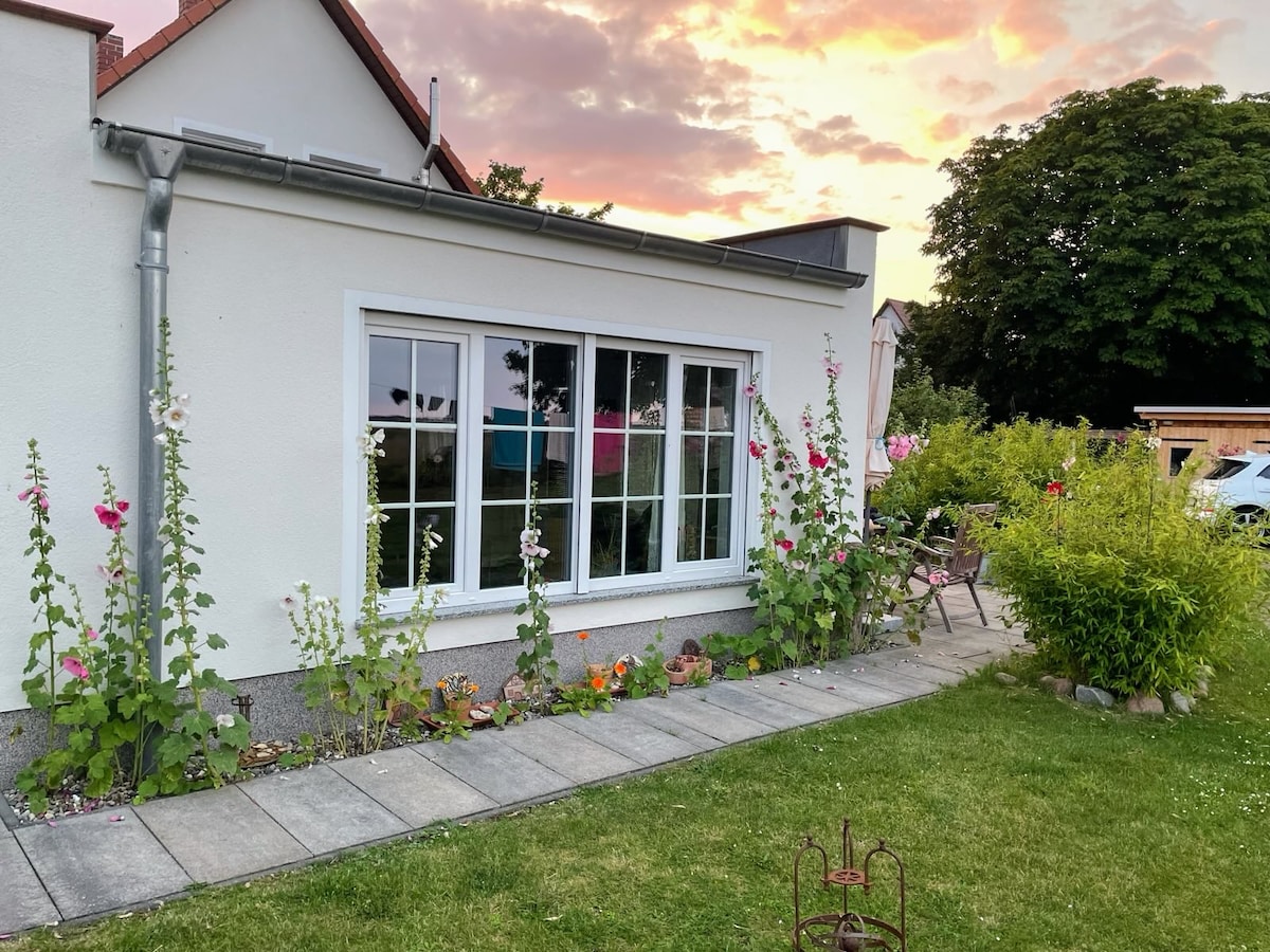 A garden area features a well-kept lawn and a pathway lined with flowerbeds. The white exterior of the building is complemented by large windows that reflect the soft colors of a sunset. Nearby, seating is positioned for outdoor relaxation.