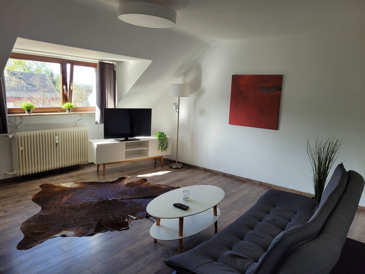 The living room features a modern design with a comfortable gray sofa and a stylish coffee table. A flat-screen TV is placed on a light-colored media console. Natural light enters through a large window, framed by gray curtains, with a decorative plant on the windowsill.