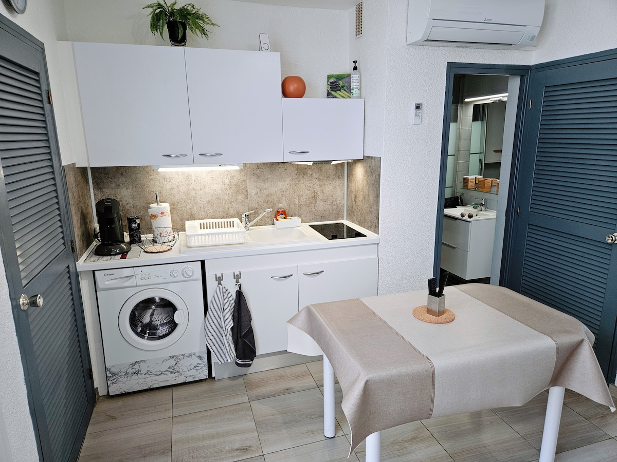A modern kitchen area is presented, featuring sleek white cabinetry and a smooth countertop. Essential appliances include a washing machine, stove, and coffee maker. A small dining table is positioned nearby, adorned with a simple centerpiece. The space benefits from natural light and neutral tile flooring.