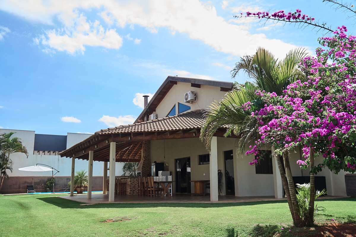 A spacious outdoor area is showcased, featuring a modern building with a thatched roof and large covered porch. Vibrant flowering plants are seen in the foreground, alongside well-maintained grass, creating a welcoming setting.