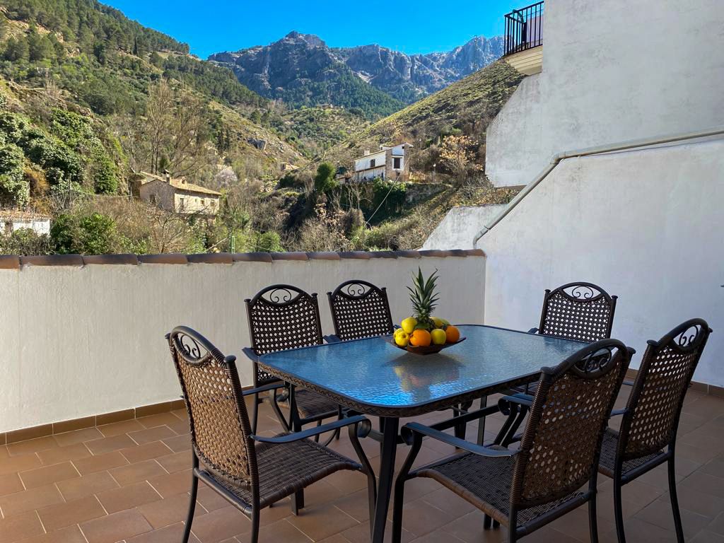 A dining area is furnished with a glass table surrounded by six wicker chairs. A centerpiece of fresh fruits is placed on the table, with a view of mountains in the distance and clear blue skies evident beyond the terrace railing.