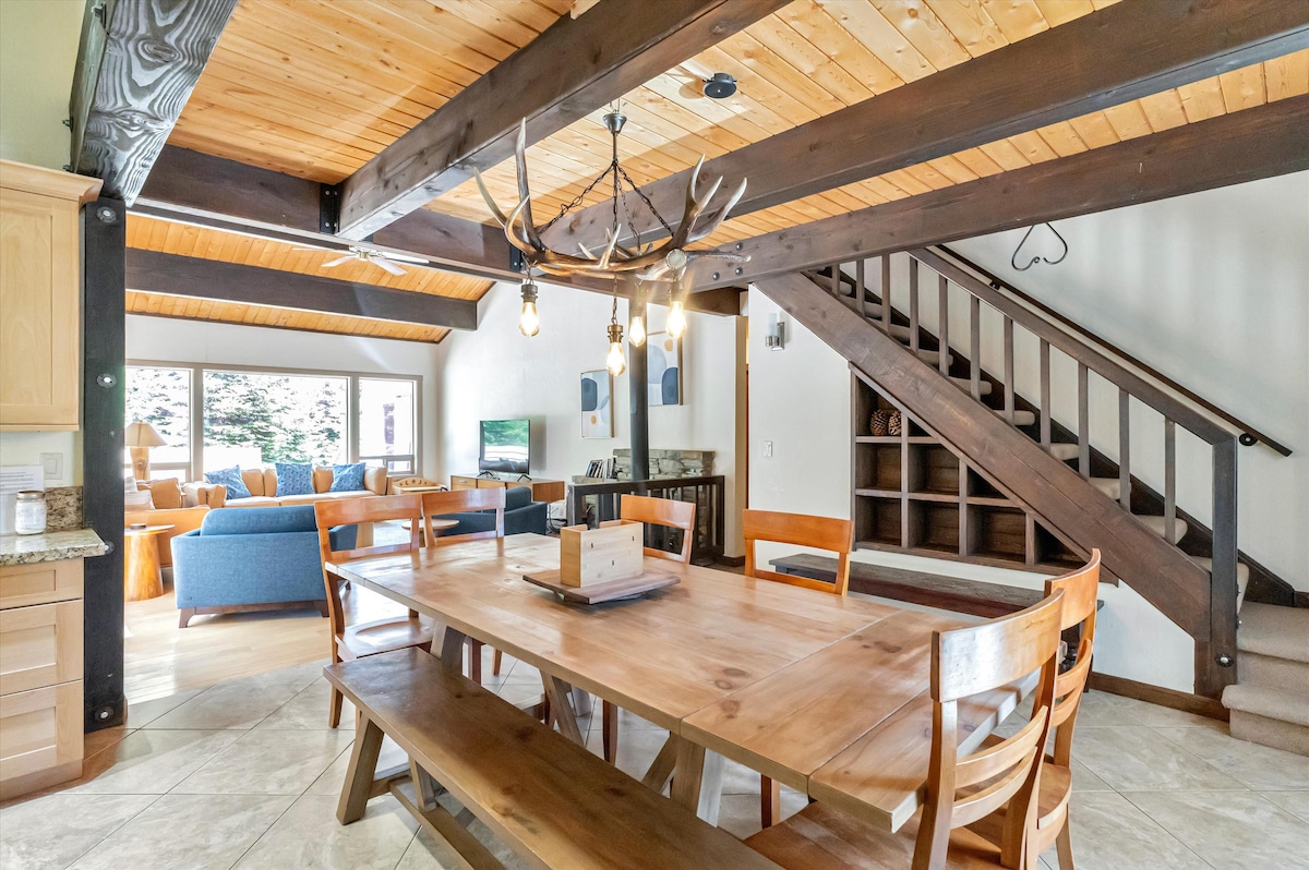A spacious dining area features a large wooden table surrounded by matching chairs, offering ample seating. Natural light filters through windows, illuminating the area. A staircase with rustic wooden railings is positioned in the background, enhancing the open and inviting layout.