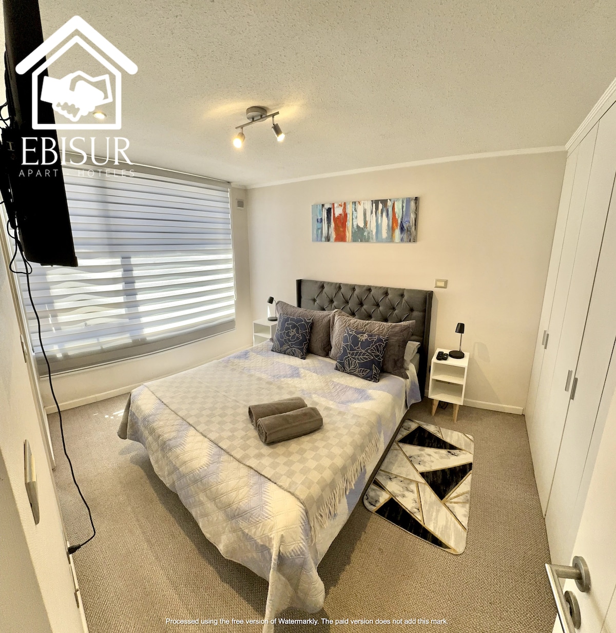 A spacious bedroom features a comfortable bed with decorative pillows and rolled towels placed on it. Nightstands are positioned on either side, supporting artistic wall decor. Soft natural light is filtered through modern window shades, creating a calm and serene atmosphere.