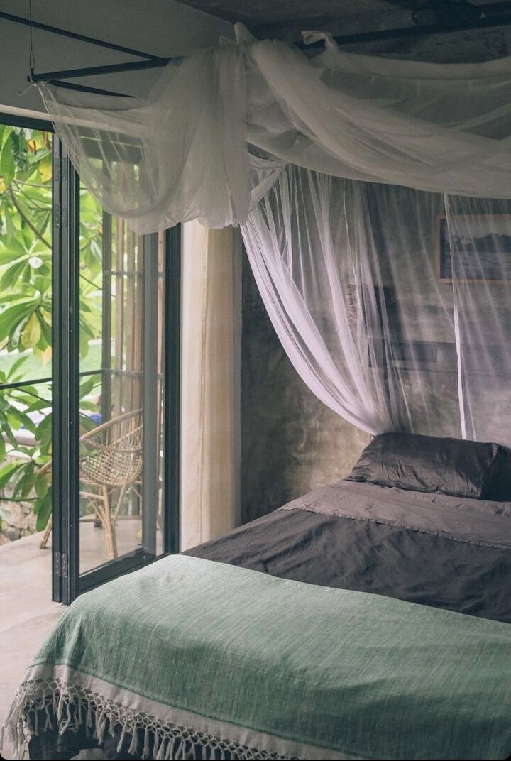 A serene room showcases a spacious bed draped with a light canopy. Natural light enters through large glass doors that provide access to a balcony, framed by lush greenery. A soft throw blanket in calming tones rests on the bed.