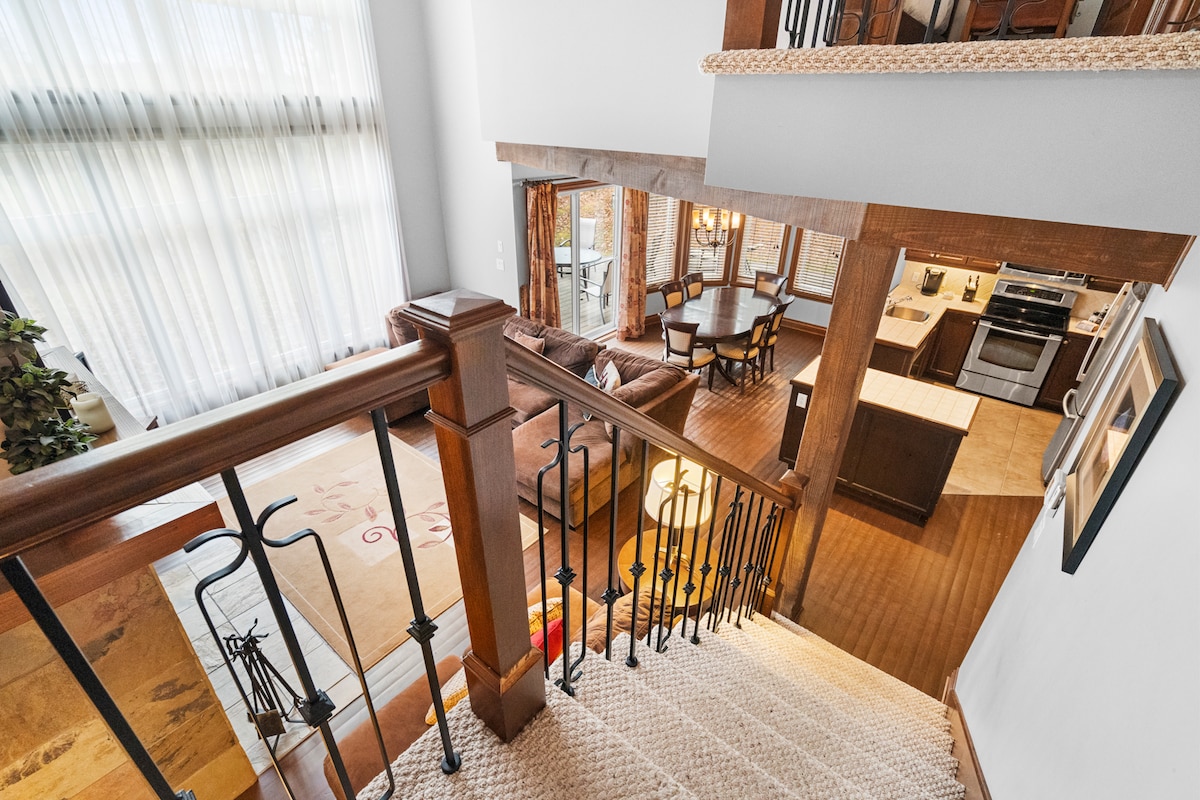An open staircase leads down to a living area with a cozy seating arrangement. Sunlight filters through large windows draped with sheer curtains, illuminating the space. A dining table and kitchen are visible through the adjacent window, creating a seamless flow between areas.