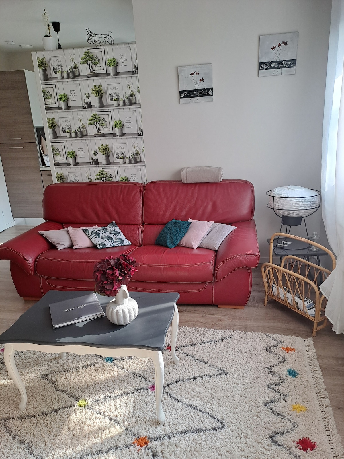 A red leather sofa is positioned in a cozy living area, complemented by decorative pillows. A small coffee table sits in front, adorned with a vase and a book. Nearby, a wicker side table and a lamp add functional details, while two framed artworks hang above.