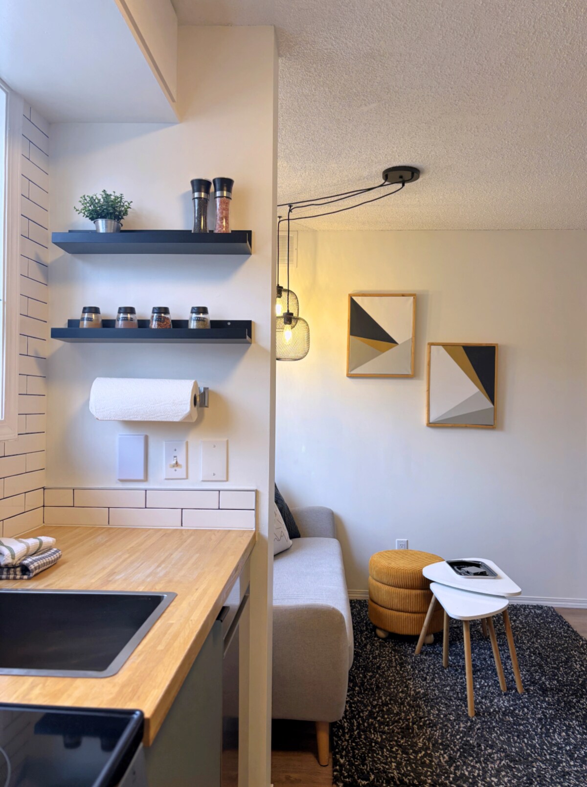 A well-organized kitchen area features light wood countertops and open shelving displaying small jars and a plant. Adjacent, a cozy living space includes a gray sofa, a small white table, and a yellow ottoman against a neutral wall adorned with framed geometric artwork.