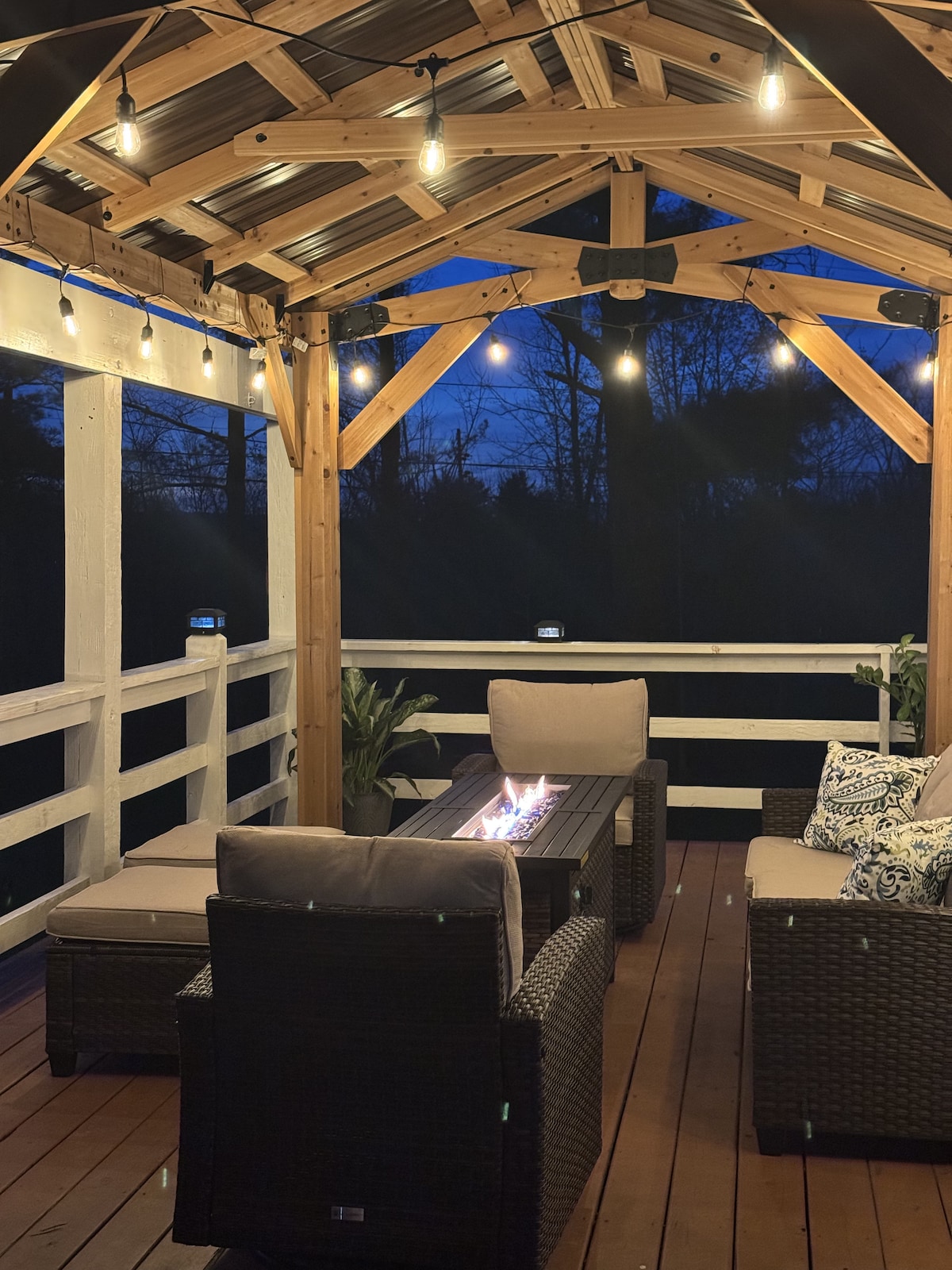 A sheltered seating area features wicker furniture with cushions arranged around a central fire table. Soft lighting from hanging bulbs creates a cozy atmosphere, while the surrounding trees are silhouetted against the dusk sky.