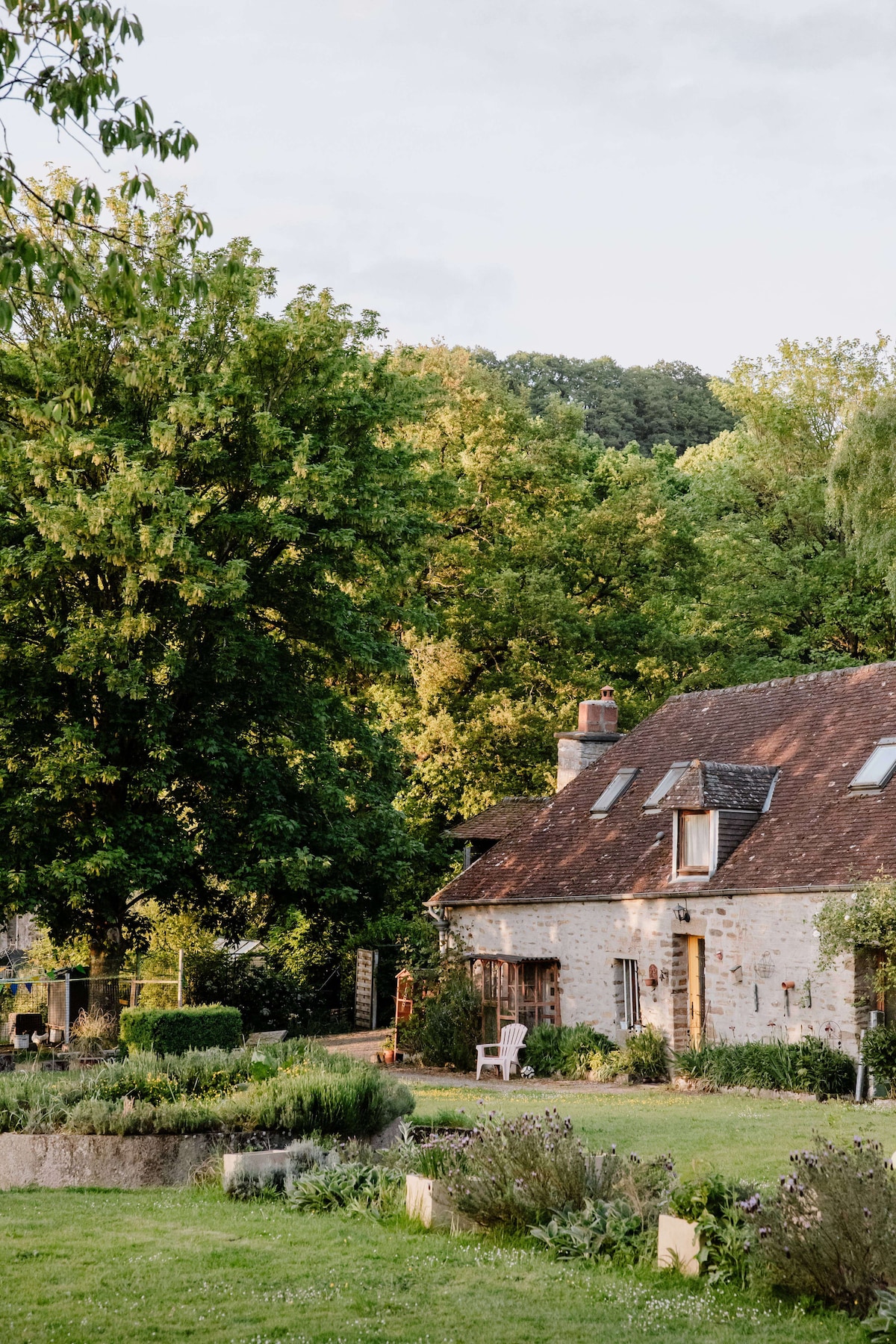 The exterior of a charming stone house is showcased, surrounded by lush greenery. A spacious lawn features neatly arranged garden beds, while large trees provide shade. The structure includes multiple windows and a distinctive sloped roof, inviting a sense of tranquility in the natural setting.