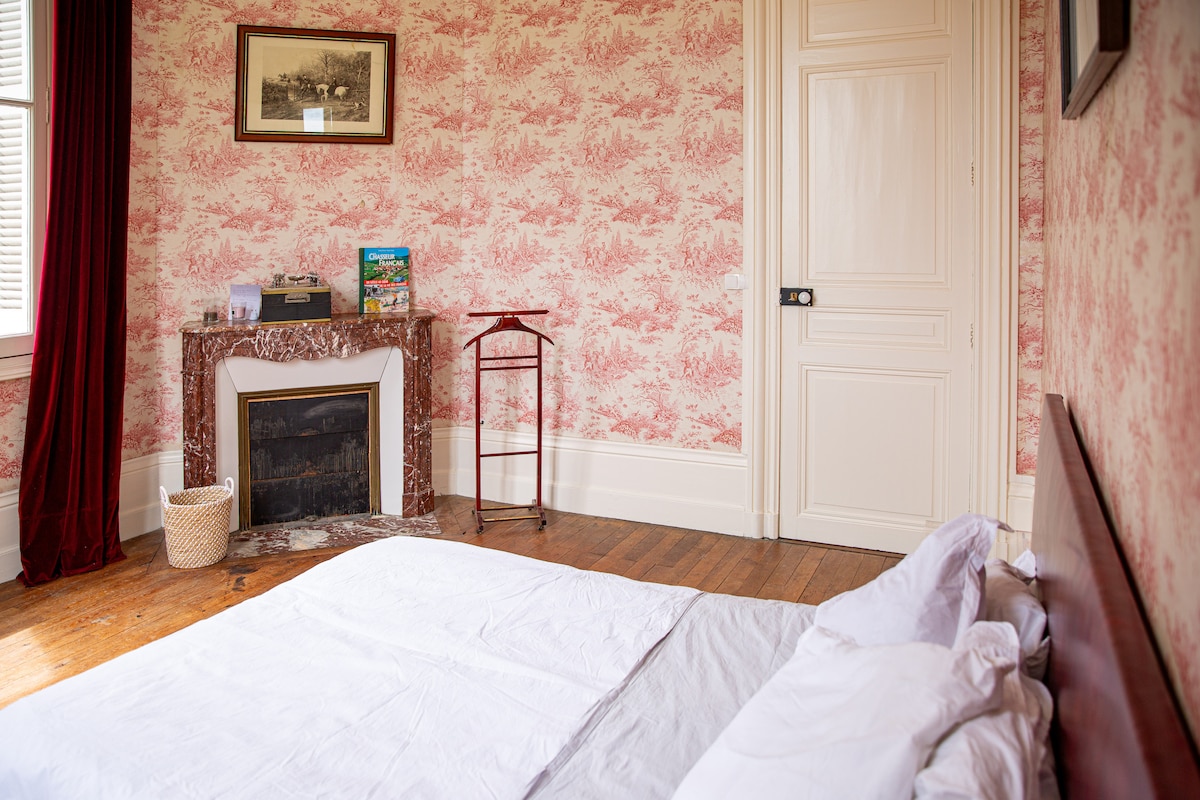 A charming room features floral-patterned wallpaper and warm wooden flooring. A spacious bed with white linens is positioned alongside a decorative fireplace made of marble, while a small reading area is provided with a wooden stand and light decor.