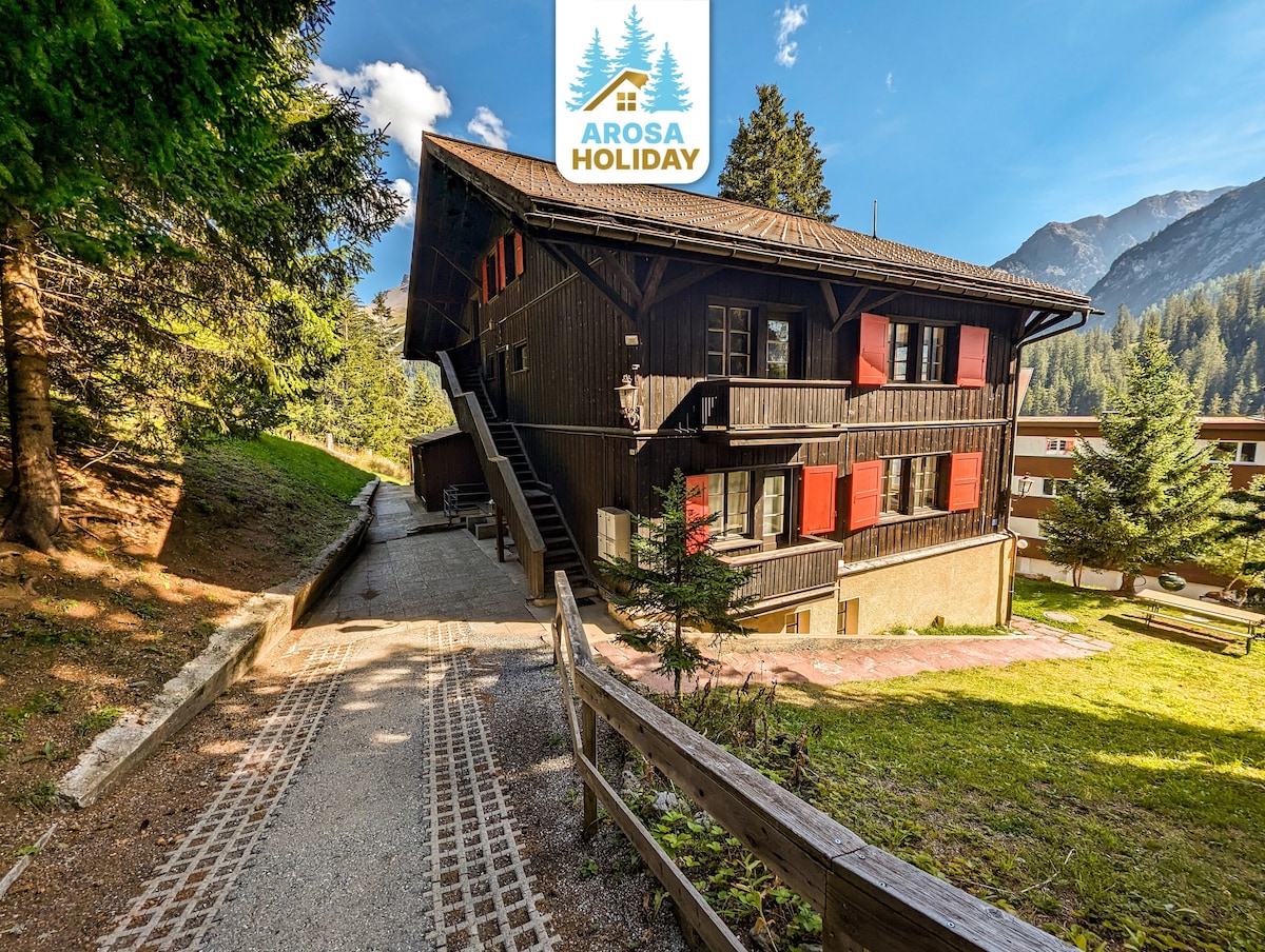 The charming wooden chalet features multiple balconies with red shutters. A pathway leads to the entrance, surrounded by lush greenery and mountain views. The building’s rustic design is complemented by the natural landscape, providing a peaceful setting for guests.