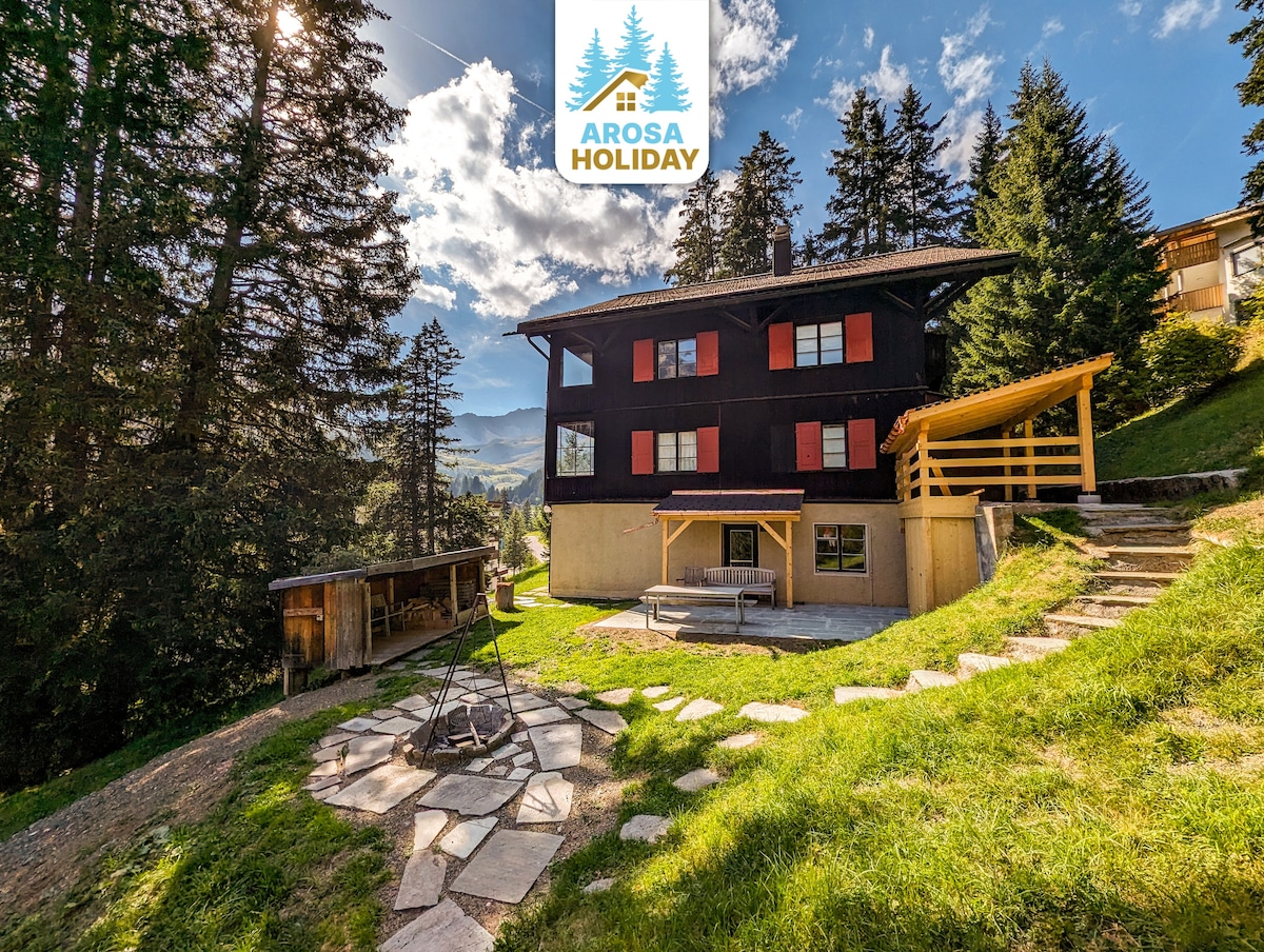 The exterior of the chalet is surrounded by lush trees, with a well-maintained garden featuring a stone pathway leading to a grilling area. The building presents a two-story structure painted in dark tones, highlighted with red shutters, under a partly cloudy sky.