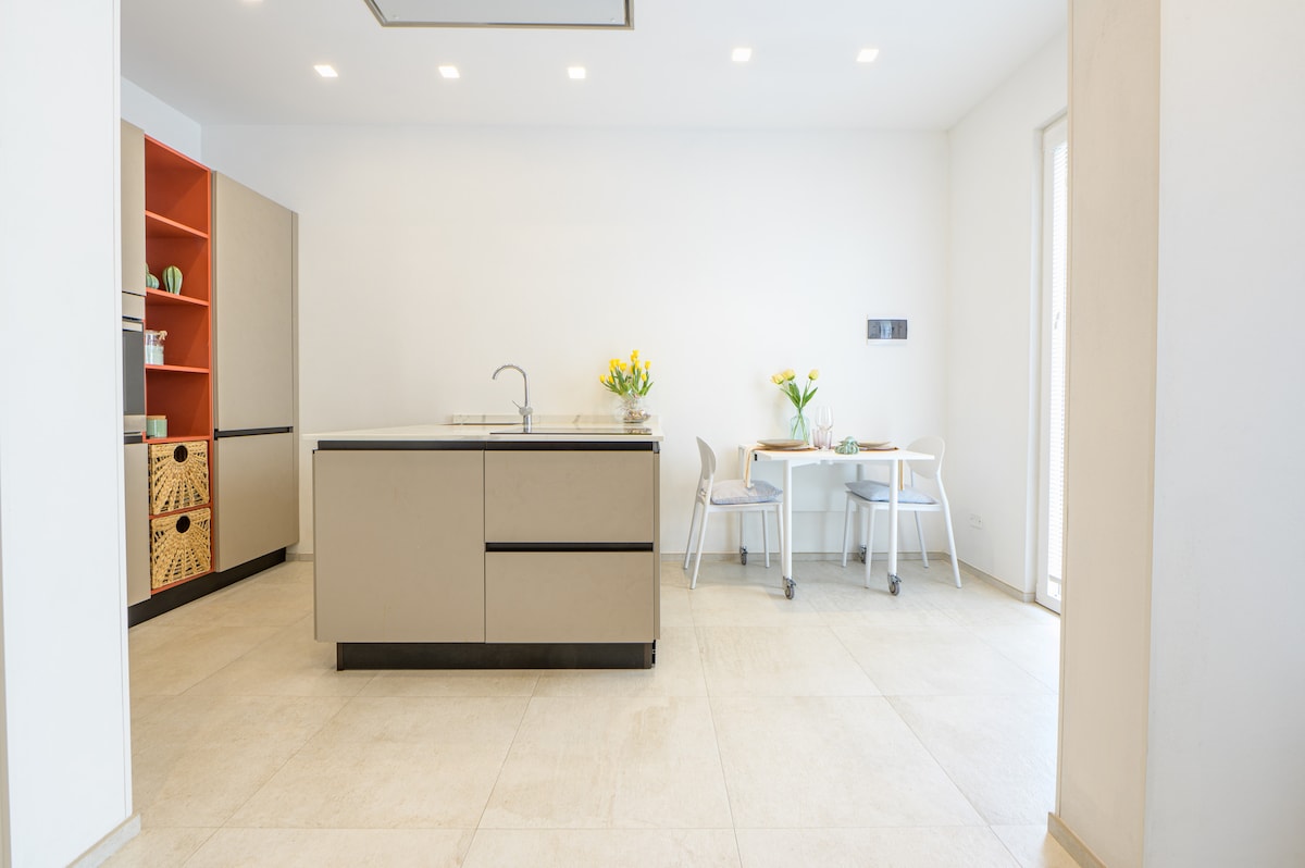 A modern kitchen area is displayed with a sleek central island and minimalist cabinetry. Natural light filters through a nearby window, illuminating a small dining table set for two. Bright flowers are arranged on the counter, adding a touch of color to the neutral palette.