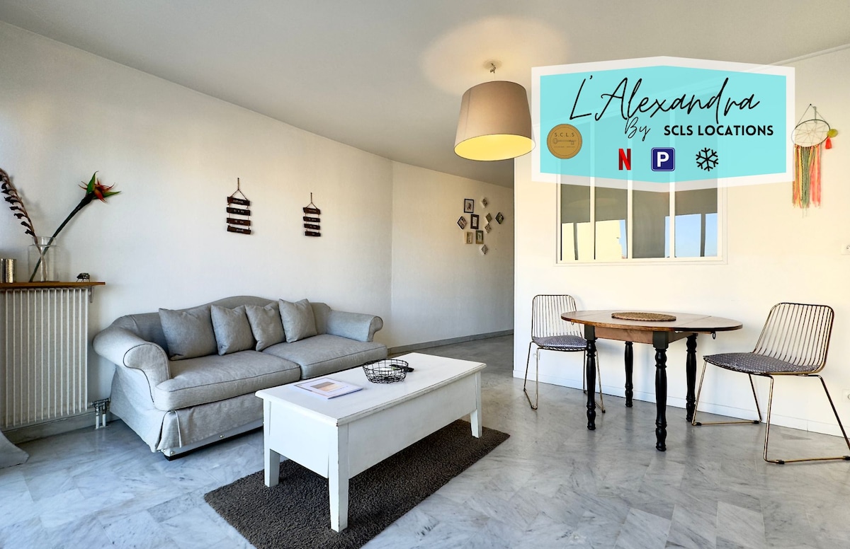 A spacious living area is presented with white walls and marble flooring. A light-colored coffee table is centered, accompanied by two wire chairs and a round dining table. Natural light is streamed in through a large window, enhancing the open and airy feel of the space.