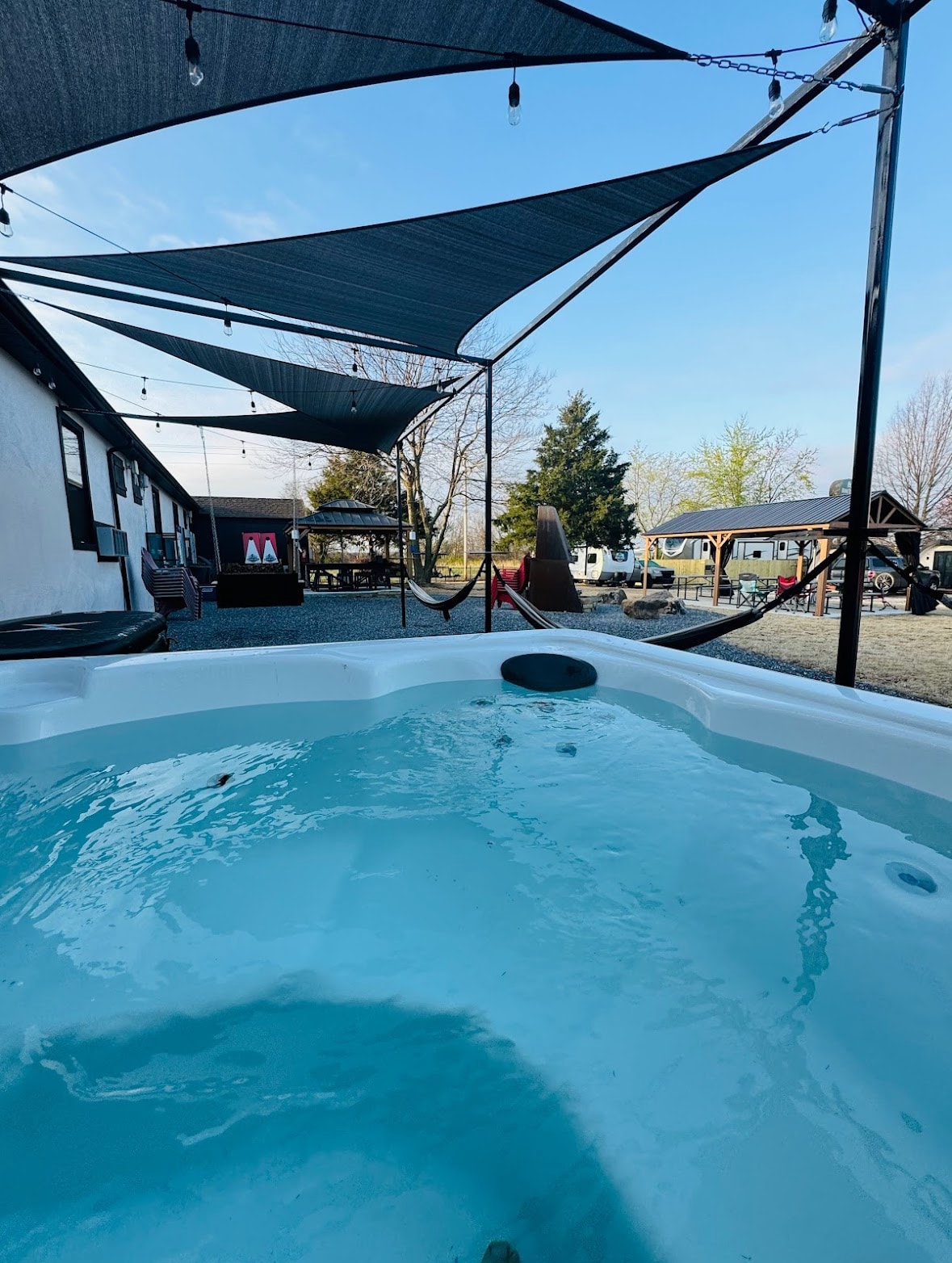 An outdoor hot tub is visible, surrounded by shaded areas created by large canopies. Hammocks are suspended nearby, offering a relaxed space for guests. The clear blue sky and trees are seen in the background, creating a serene outdoor setting.