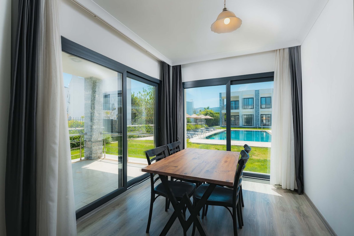 A bright dining area is illuminated by natural light from large glass doors, which offer a view of a serene outdoor space featuring a pool and well-maintained greenery. A wooden dining table is positioned in the center, complemented by black chairs.