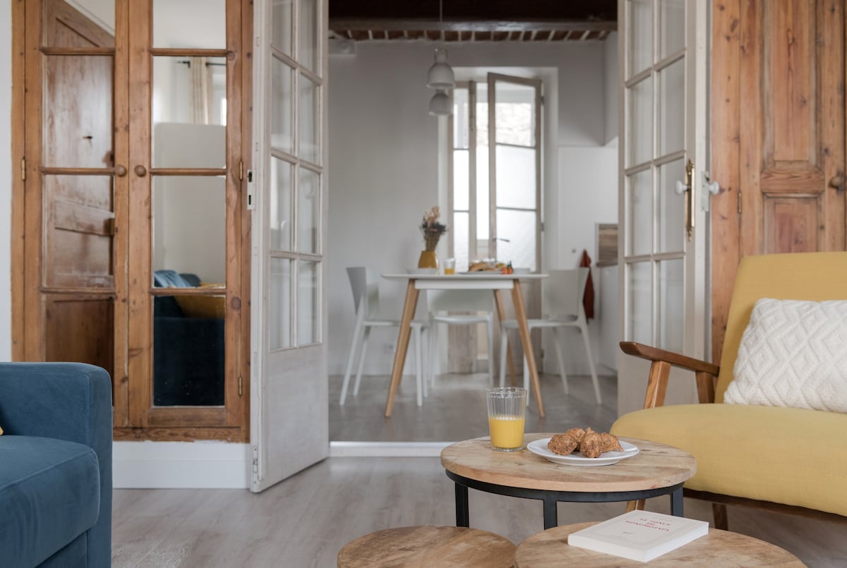 A cozy living space is framed by glass-paneled doors, leading to a bright dining area with a wooden table set for breakfast. Two small tables, one holding a glass of juice and pastries, are positioned in the foreground amidst a blend of modern and rustic furniture.