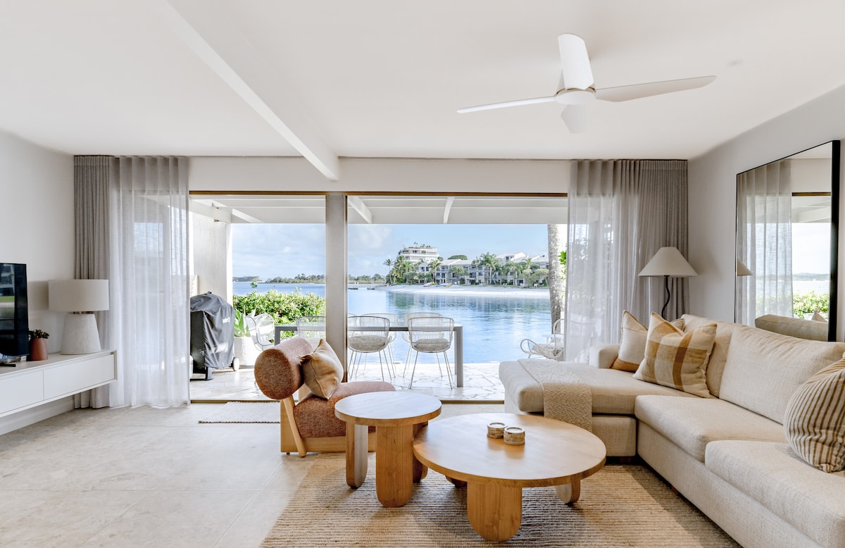 A light-filled living area features a comfortable sectional sofa and two round wooden tables. Large sliding glass doors open to scenic water views, with outdoor seating visible on a patio. A ceiling fan circulates air, enhancing the open and airy atmosphere.