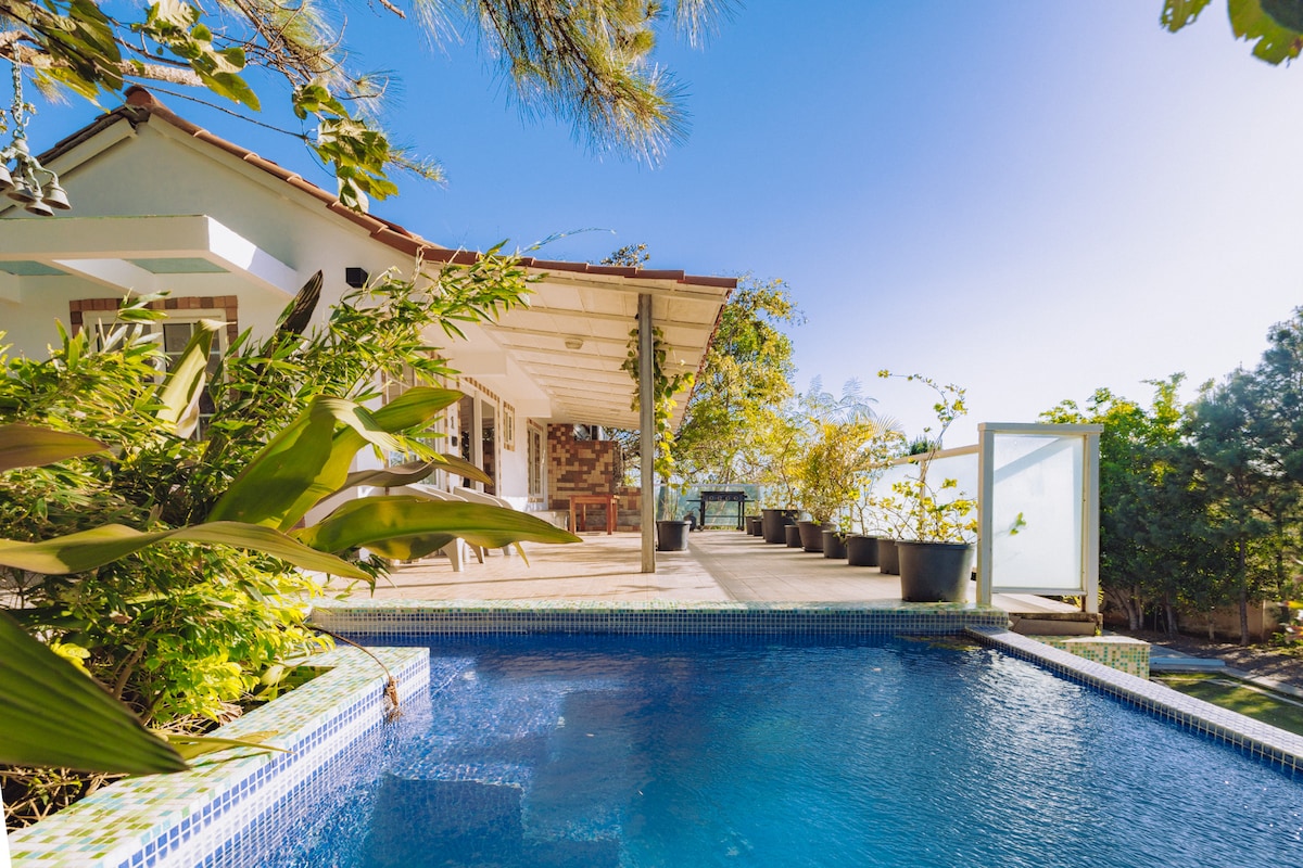 A serene outdoor terrace features a refreshing swimming pool, surrounded by lush greenery and potted plants. The house's entrance, accentuated by wooden beams, opens to a spacious area with bright natural light. Clear blue skies complete the tranquil setting.