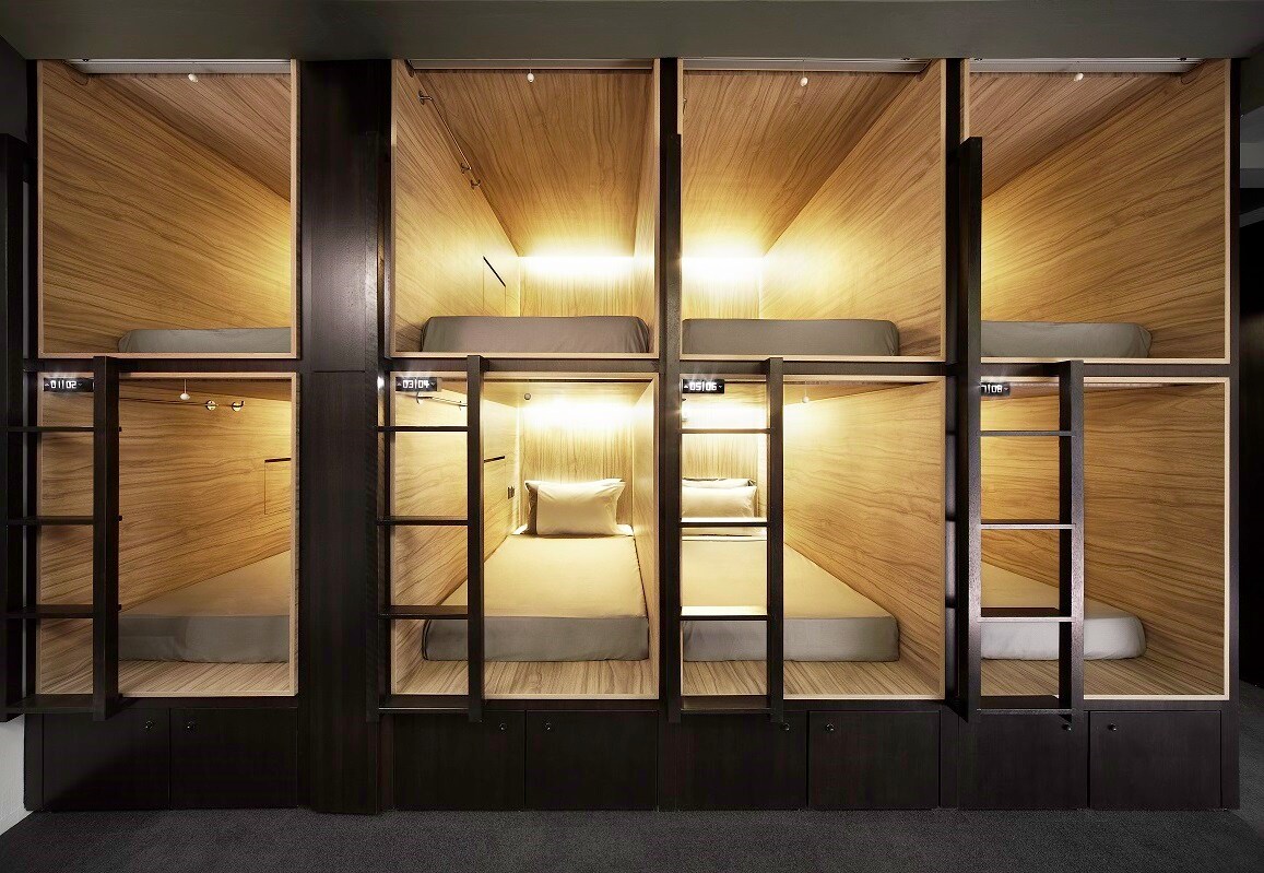 The image displays a series of modern capsule pods arranged in a shared space, each featuring a compact sleeping area with a cushioned mattress and soft lighting. Wooden paneling creates a warm aesthetic, while ladders provide access to the upper pods.