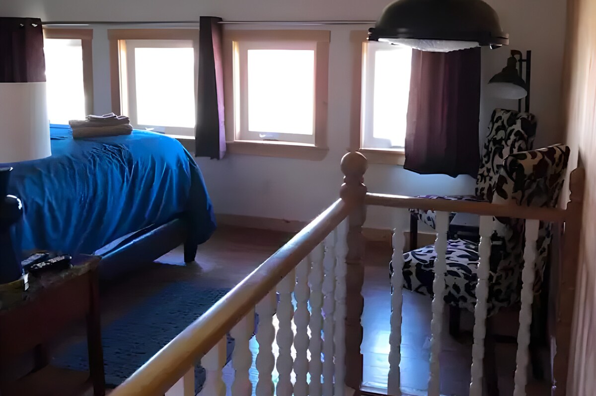 A bright bedroom is framed by multiple windows, allowing natural light to fill the space. A spacious bed covered in blue linens is visible, accompanied by a patterned armchair in the corner. Soft curtains frame the windows, enhancing the tranquil atmosphere of the room.