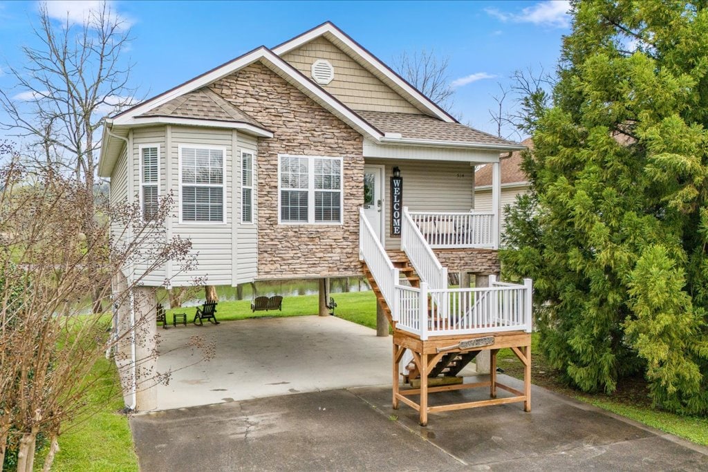 A spacious elevated home features a stone facade and large windows, allowing natural light to fill the interior. A covered porch with steps leads to the entry. The surrounding area is landscaped with green grass and trees, providing a peaceful environment.