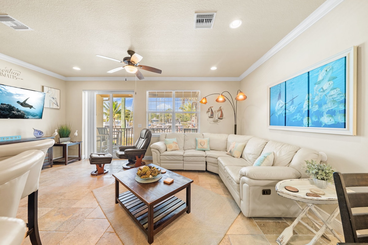 A bright living area features a large sectional sofa in a curved arrangement. A coffee table is positioned in front, surrounded by casual seating. Natural light enters through multiple windows, illuminating the space. Decorative wall art and a ceiling fan add to the homey feel.