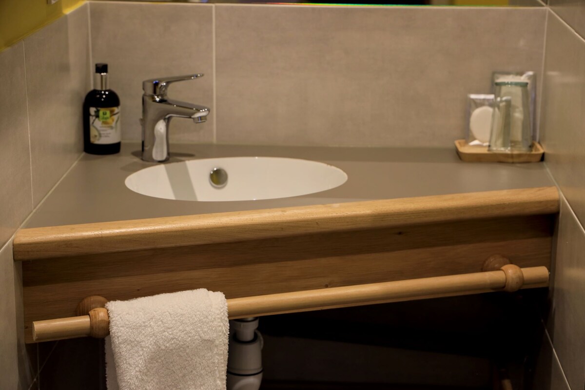 A modern bathroom sink area is displayed, featuring a circular white sink set into a gray countertop. Below the counter, a wooden towel rack is visible, holding a white towel. A stylish faucet and various toiletries are positioned nearby.