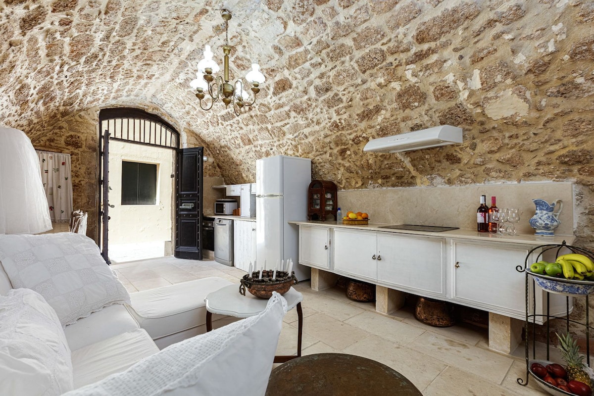 An inviting kitchen area features a stone arched ceiling, with light natural wood cabinetry and a white refrigerator. A comfortable sofa is positioned nearby, alongside a small dining table with fresh fruit. Double doors lead to the outside, enhancing the bright and airy feel.