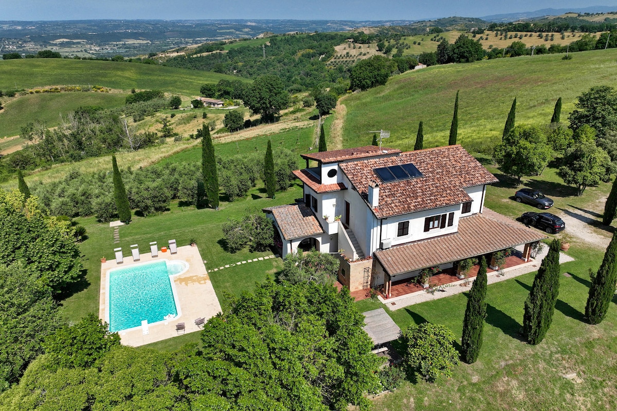 An aerial view of the villa showcases its three-story architecture set among lush greenery. A private swimming pool and surrounding lounge area are visible, framed by manicured lawns and tall cypress trees. A scenic landscape of rolling hills extends into the distance.