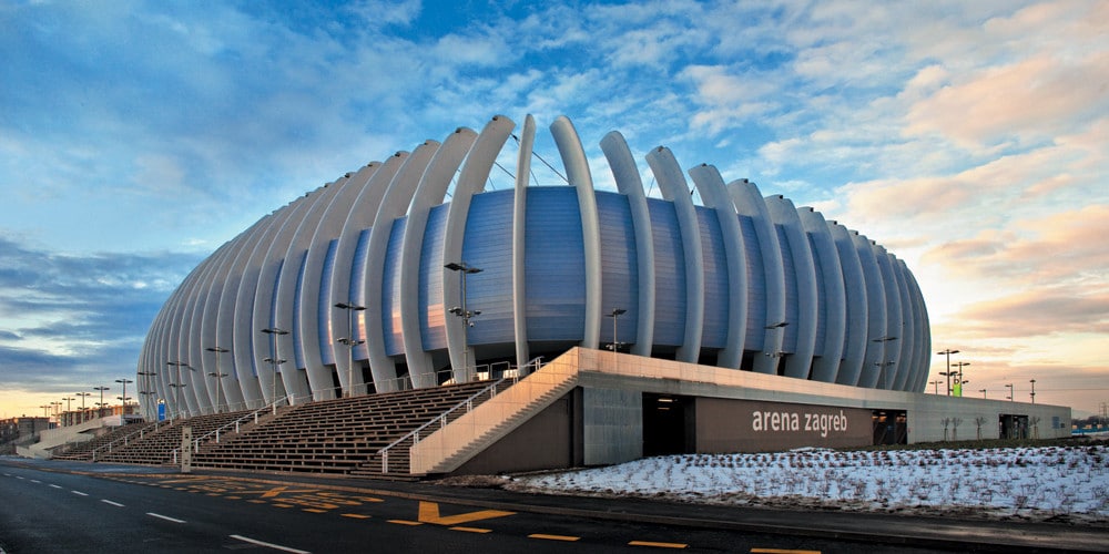 A modern stadium is prominently featured, characterized by its unique exterior design and large, flowing lines. The structure is set against a sky that transitions from light to dark, while the surrounding area includes a wide path and gentle snow cover.