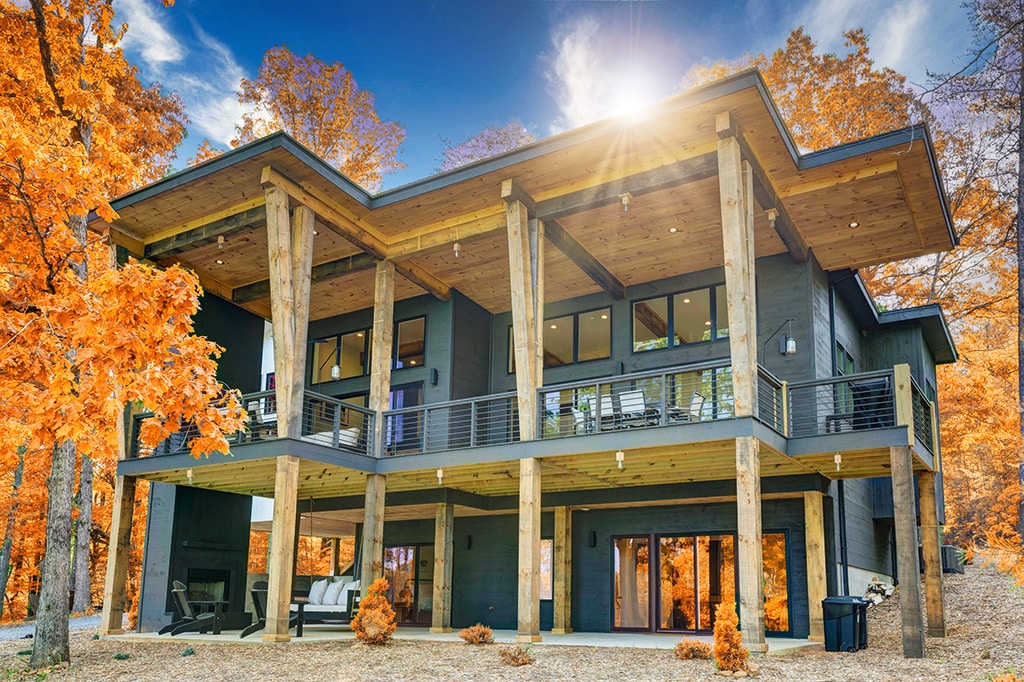 A modern cabin is showcased, featuring an expansive deck supported by tall wooden columns. The structure is surrounded by vibrant autumn foliage, with large windows offering views of the outdoors. Comfortable seating is arranged on the patio, inviting relaxation amid nature.