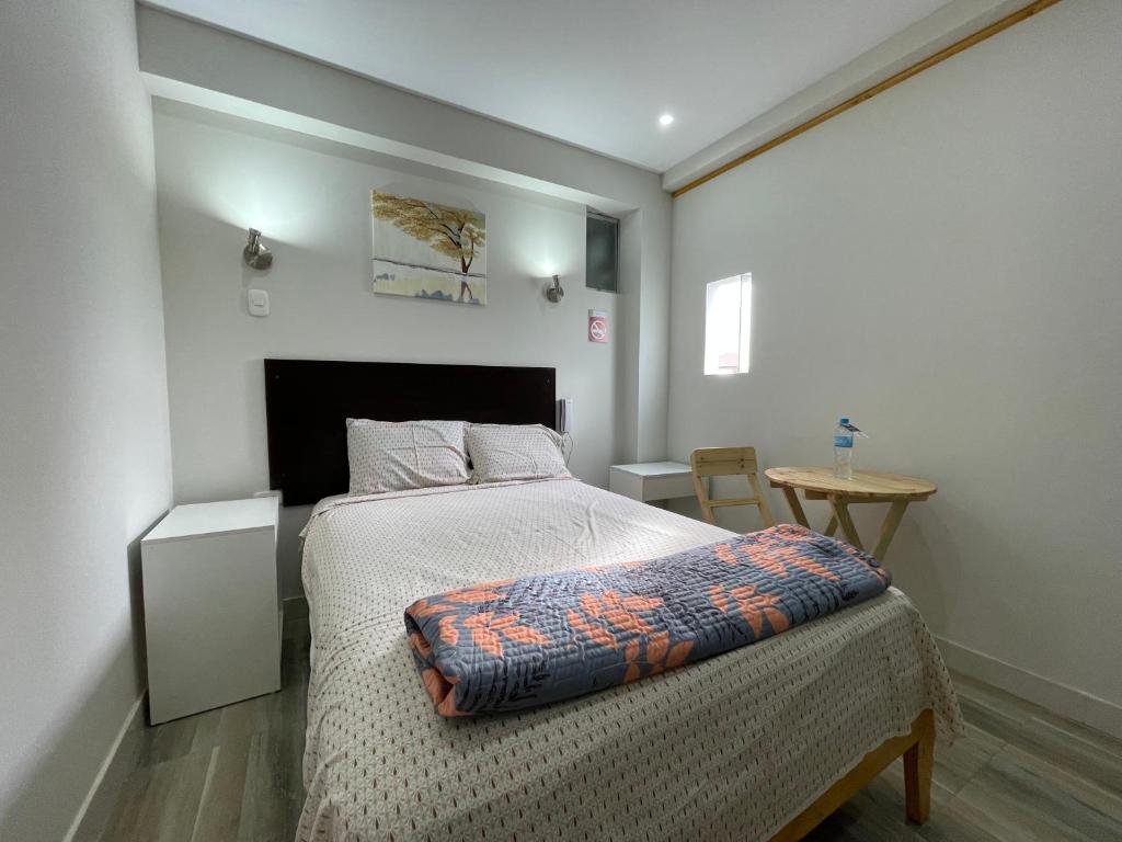 A comfortable sleeping area is presented with a spacious bed dressed in light-colored linens. Beside the bed, a small table and a wooden chair are arranged. Natural light enters through a small window, and an artwork depicting a tree is displayed on the wall.