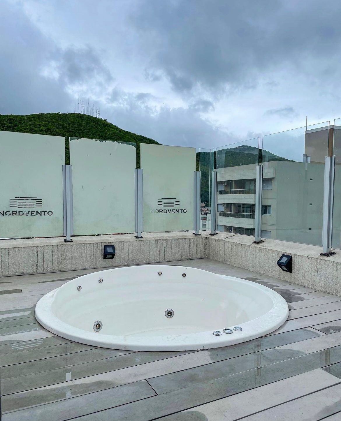 A spacious outdoor area features a round hot tub surrounded by glass panels, with views of the surrounding landscape. The decking has a wood-like finish, and lighting fixtures are installed at the base, enhancing the overall space.