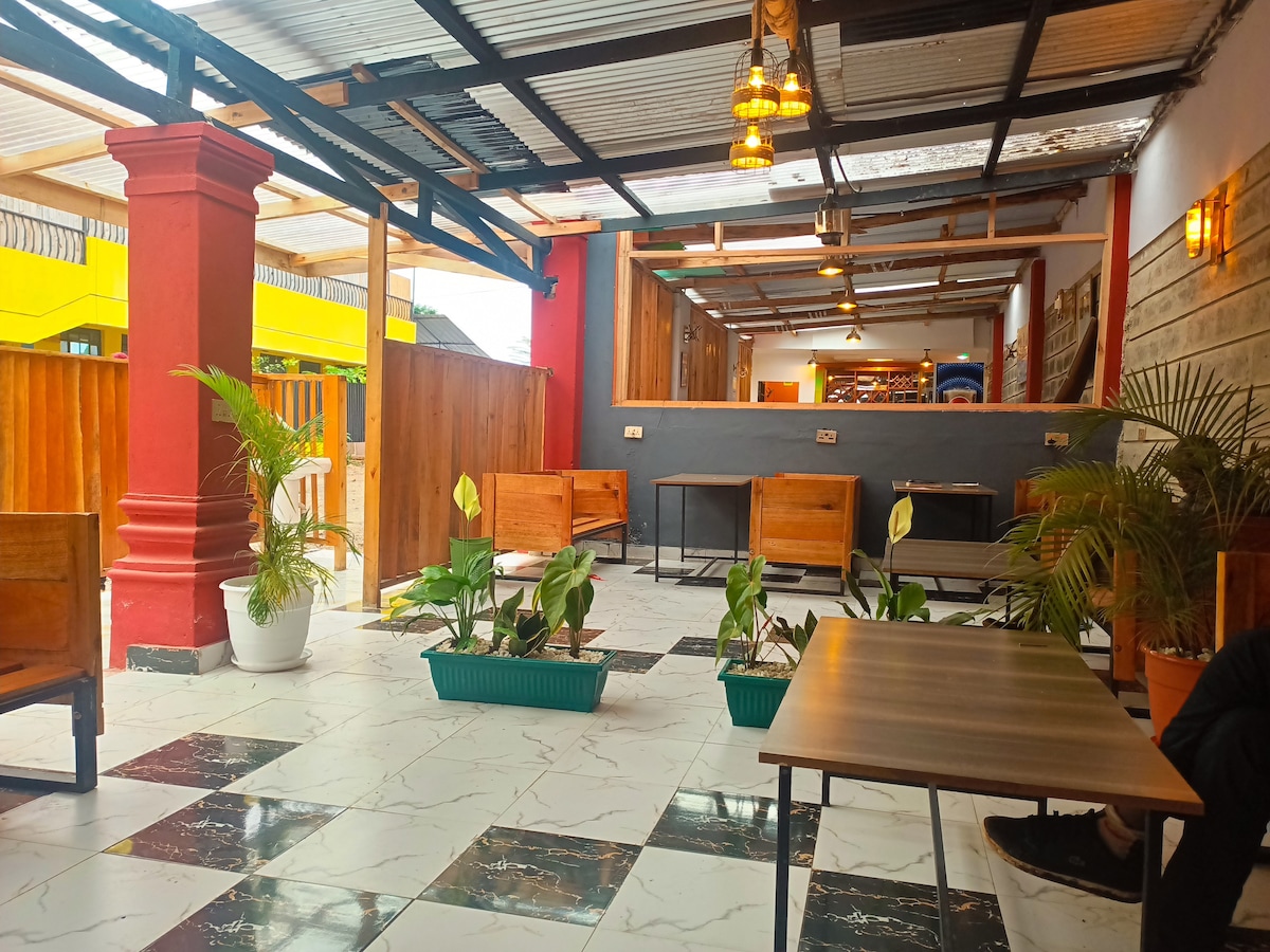An open dining area features a combination of wooden seating and tables nestled among decorative plants. The floor displays a bold black and white pattern. Warm lighting fixtures create a welcoming ambiance against the vibrant walls.